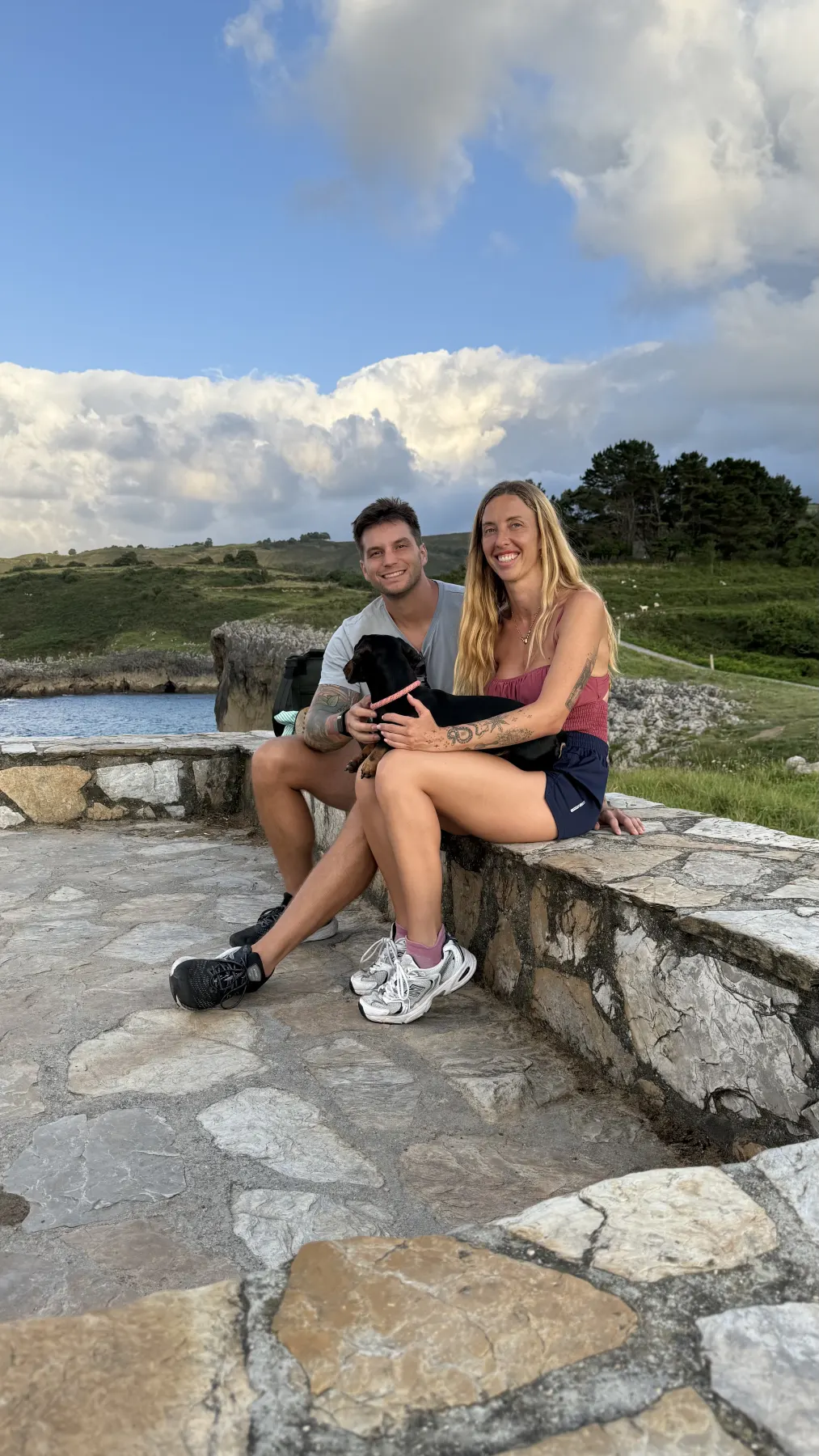 Los dos sentados en un muro de piedra junto al mar, con un perro en brazos.